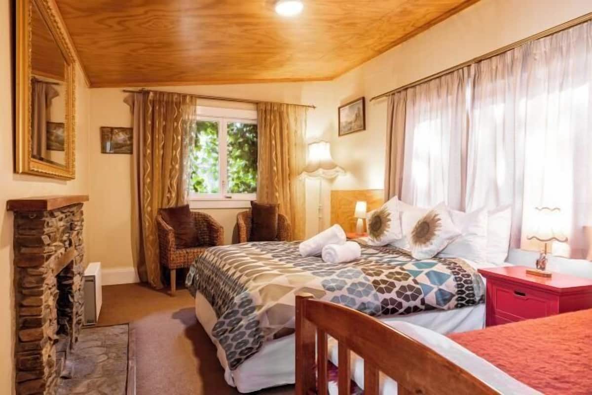 Une chambre avec une cheminée en pierre, un lit avec une literie à motifs, une table de chevet avec une lampe et une fenêtre avec des rideaux.