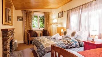 Une chambre avec une cheminée en pierre, un lit avec une literie à motifs, une table de chevet avec une lampe et une fenêtre avec des rideaux.