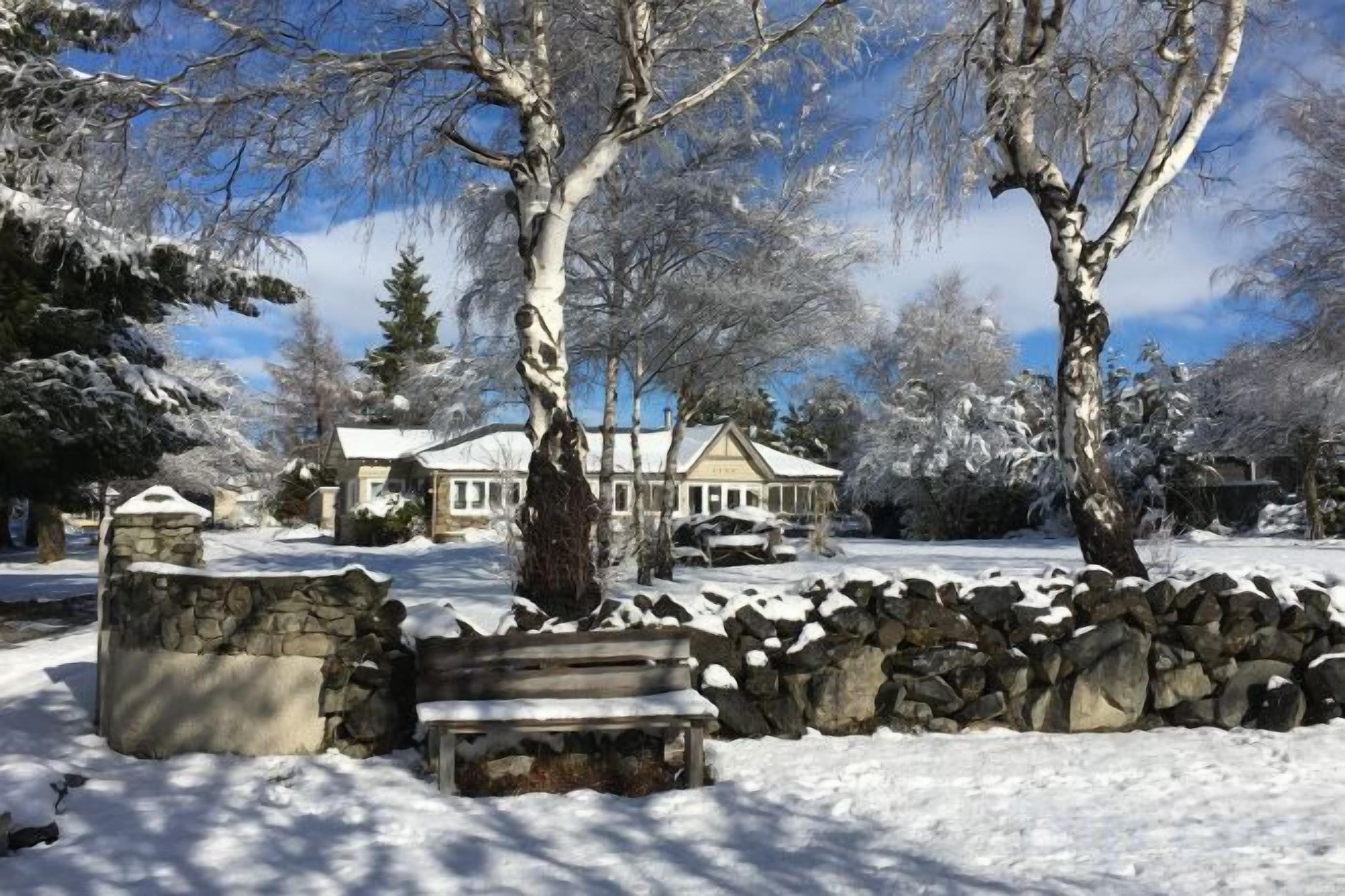 A snow-covered landscape with a stone wall, a bench, and a house in the background.