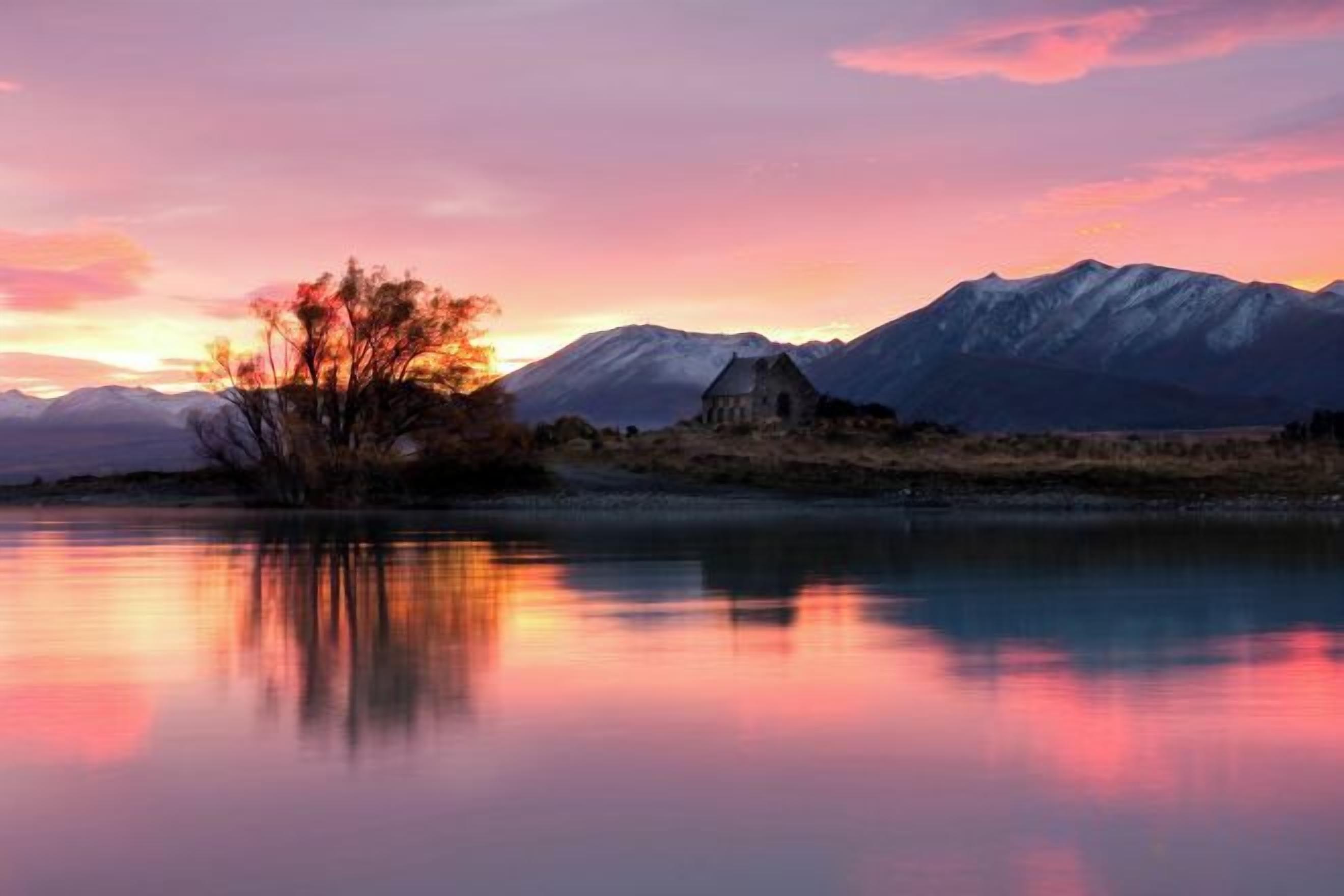 A tranquil lakeside scene with a mountain range in the background and a small building near the water's edge.