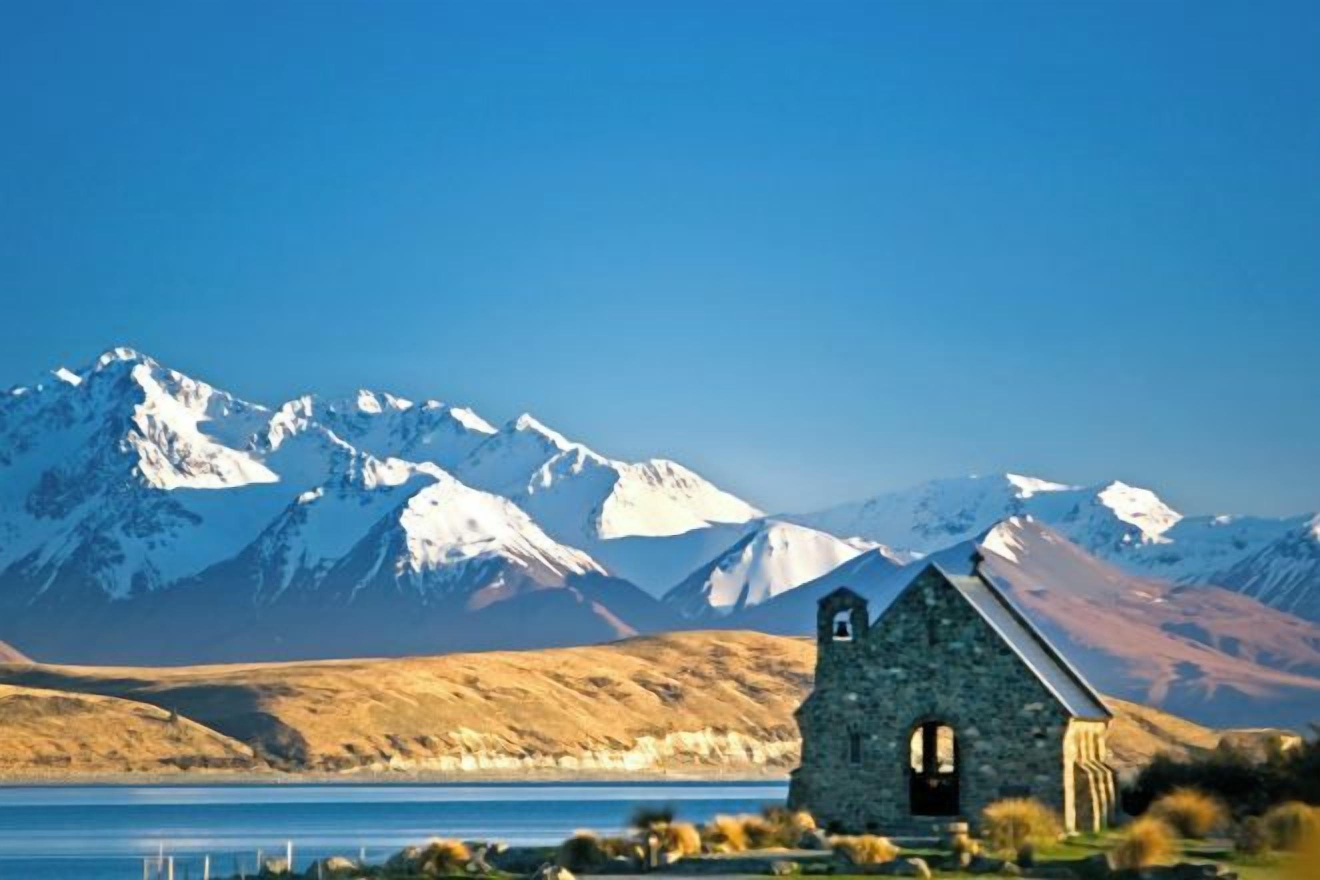 A stone church with a bell tower, surrounded by a lake and snow-capped mountains in the background.