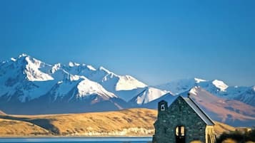 A stone church with a bell tower, surrounded by a lake and snow-capped mountains in the background.