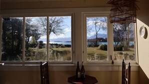 Ein Zimmer mit großen Fenstern und Blick auf einen See und Berge. Ein Holzsessel und ein kleiner Tisch mit Flaschen sind vorhanden.