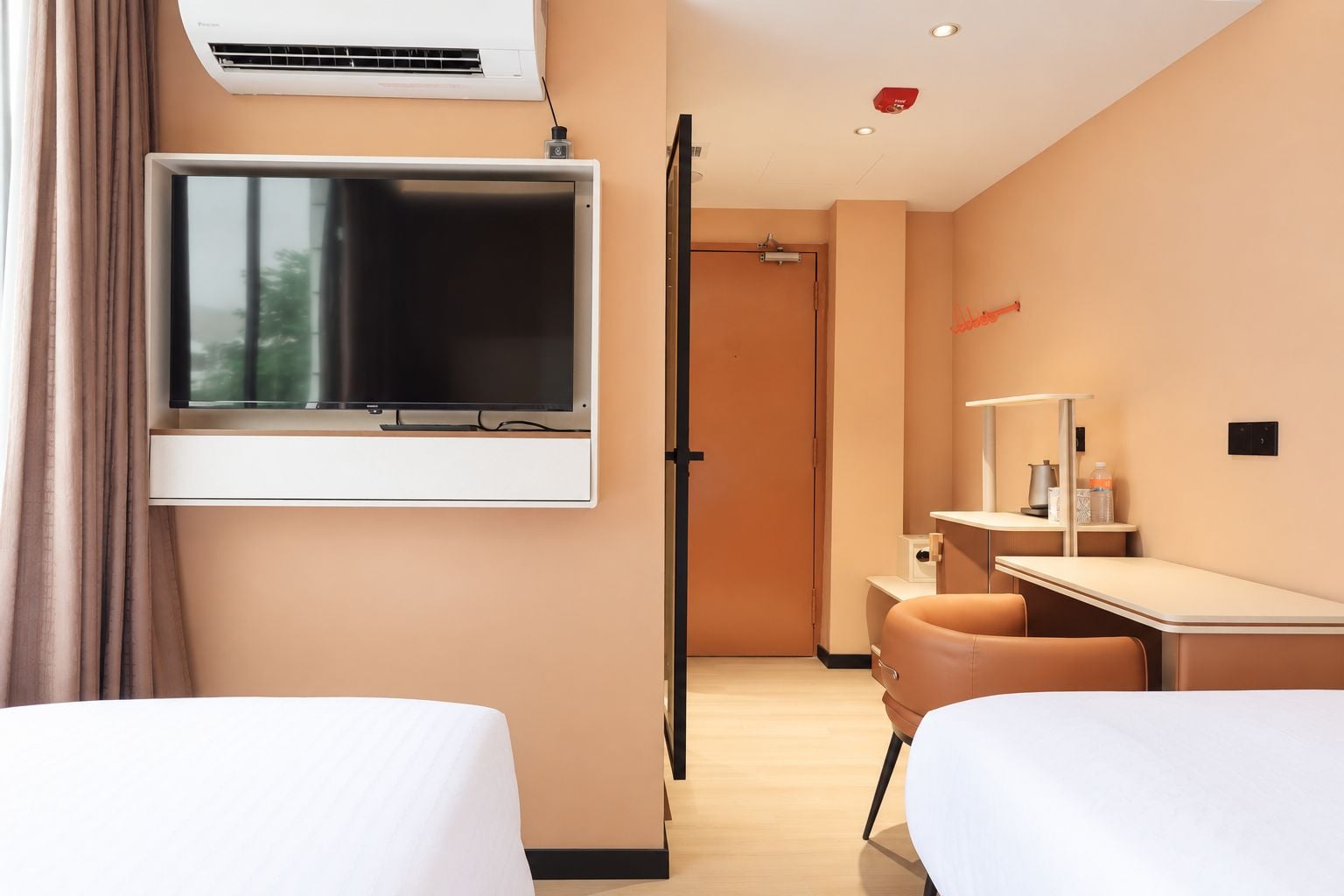 A hotel room with a bed, a desk, a chair, a TV mounted on the wall, and an air conditioning unit.