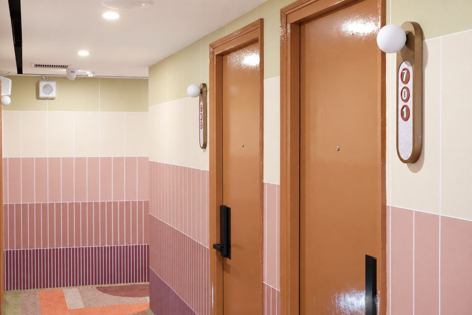 A hotel room with two wooden doors, a tiled wall, a small rug, and a wall-mounted light fixture.