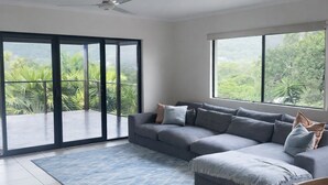Amplia sala de estar con un sofá seccional gris, alfombra azul y grandes ventanales con vista a follaje.