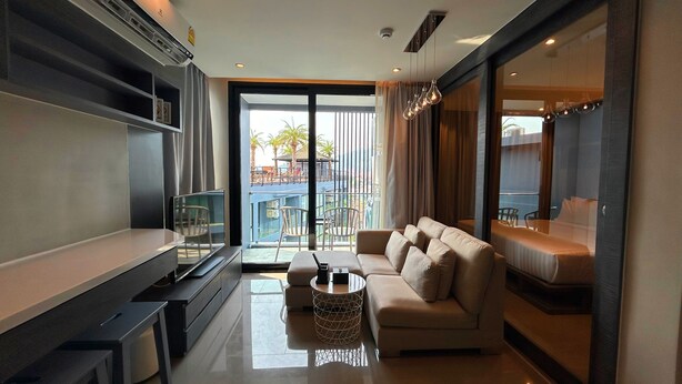 A modern living room with a sofa, a coffee table, and a TV. There is a balcony with outdoor furniture visible through the glass doors. - 1BR with Rooftop Pool & Gym  C191 (Kamala)