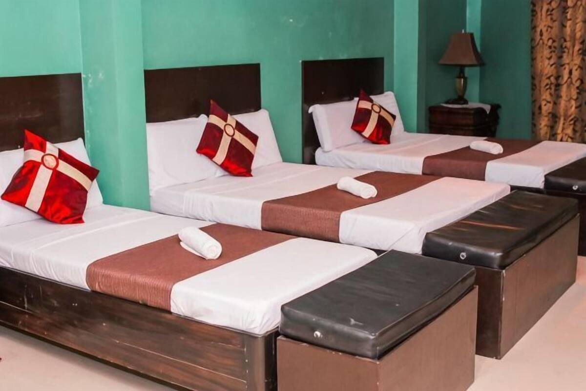 Ein Hotelzimmer mit drei Betten, jeweils mit einem hölzernen Kopfteil, weißen Leinen und roten und weißen Kissen. Ein Nachttisch mit Lampe und ein Vorhang sind ebenfalls vorhanden.
