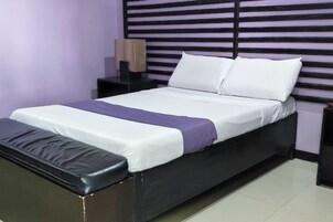 Una cama bien hecha con ropa de cama blanca y morada, un cabecero negro y un banco a juego al pie de la cama.
