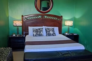 Una cama bien hecha con almohadillas estampadas, cabecera de madera, dos lámparas de noche y un espejo en la pared.