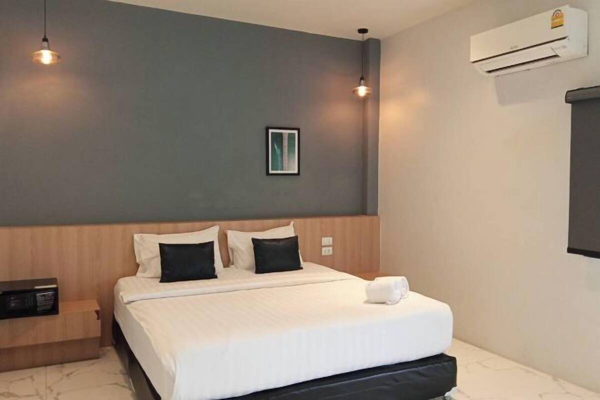 A hotel room with a bed, bedside table, wall-mounted air conditioner, and a framed picture on the wall.