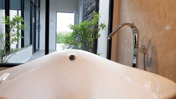 Un baño moderno con un lavamanos ovalado grande, un grifo y vistas a un pasillo con plantas y una puerta.