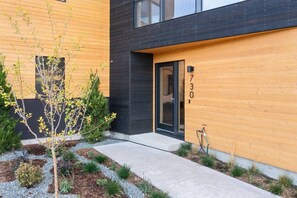 Un bâtiment moderne avec une façade en bois, un chemin en béton et un petit arbre au premier plan.