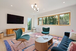 Un salon moderne doté d’un téléviseur à écran plat, de deux fauteuils, d’un canapé, d’une table basse et d’une grande fenêtre offrant une vue sur les arbres.