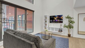 A modern living room with a grey sofa, a glass coffee table, a TV mounted on the wall, and a potted plant. - Cozy 1-bedroom loft ~ Playhouse Square ~ Fitness, BBQ, Fire pit, business center (Cleveland)