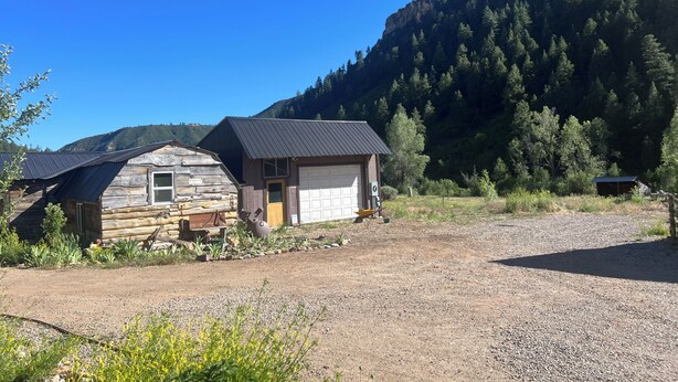 En rustik hytte med garage, omgivet af en grusvej og en skovklædt skråning.