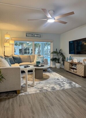A modern living room with a sofa, coffee table, and TV. There's a ceiling fan, a welcome sign, and a view of palm trees outside. - Cozy 1-bedroom condo across street from beach in terrific North Redington Beach (North Redington Beach)