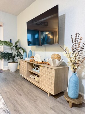 A modern living room with a wooden cabinet, a flat-screen TV mounted on the wall, and decorative plants. - Cozy 1-bedroom condo across street from beach in terrific North Redington Beach (North Redington Beach)