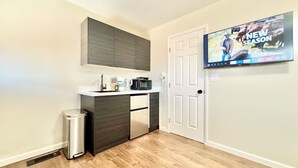 A compact kitchen with a microwave, coffee maker, and a trash can. A television mounted on the wall displays a program about the new season. - Remodeled Spanish Bungalow Suite • Short Walk to SJSU + Parking (San Jose)