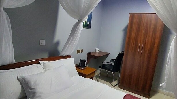 A hotel room with a bed, a wooden wardrobe, a desk with a chair, and a small table with a coffee pot. - ALLENS HOTEL (Kapsabet)