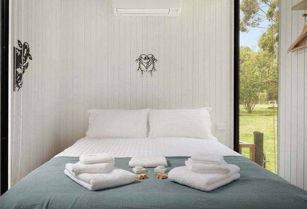 A neatly made bed with white linens and towels, a window with a view of greenery, and a wall-mounted air conditioning unit.
