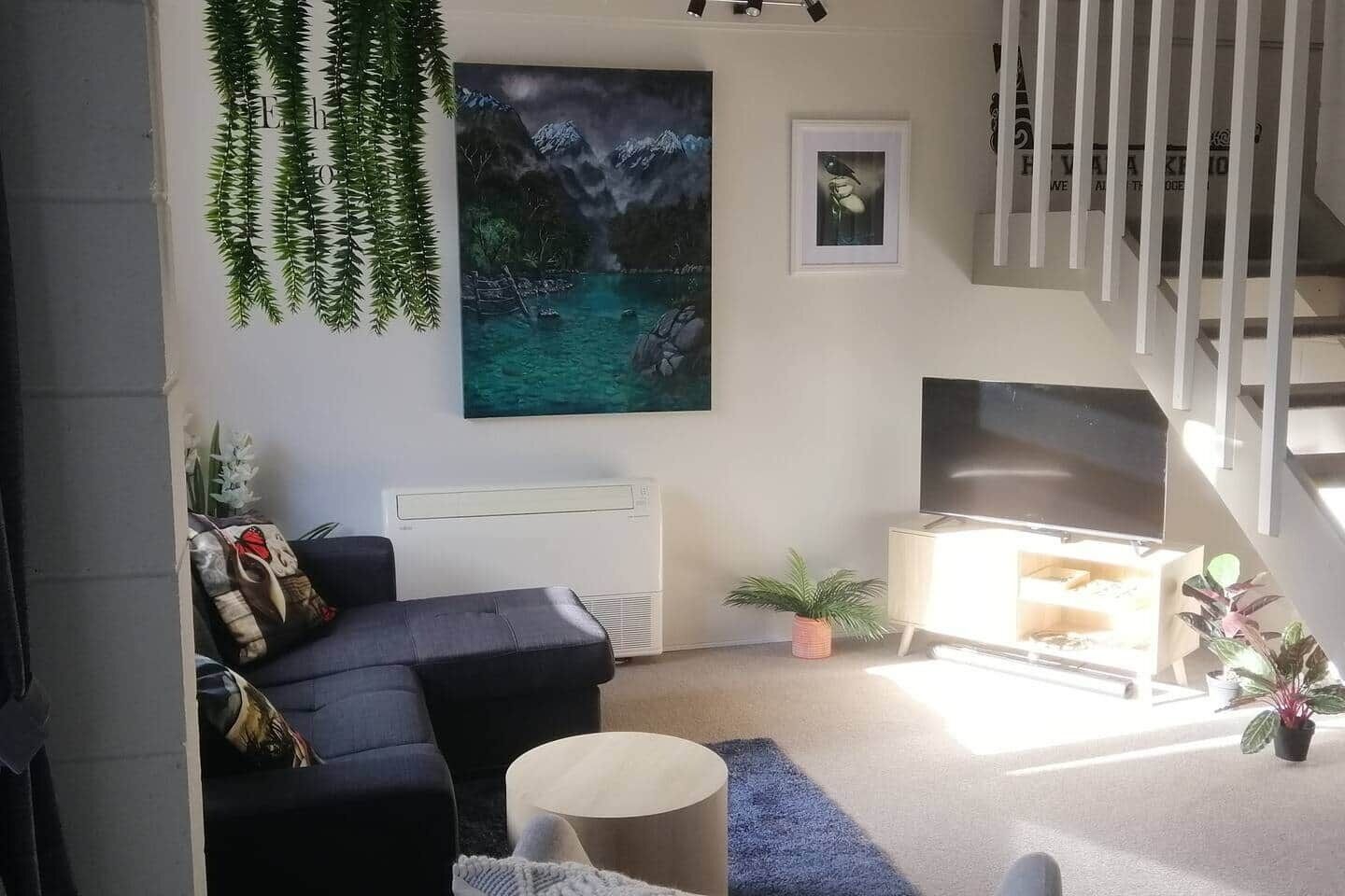 Ein modernes Wohnzimmer mit einem dunklen Sofa, einem runden Couchtisch und einem großen Gemälde einer Berglandschaft. Eine Treppe mit Geländer führt zu einer oberen Ebene.