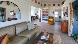Un salon spacieux avec un canapé beige, une table basse sur laquelle sont posés des livres, un ventilateur de plafond et une bibliothèque remplie de livres.