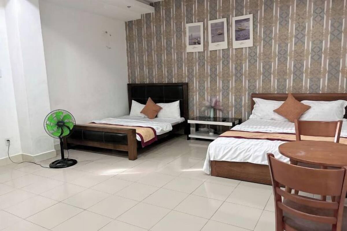A hotel room with two beds, a bedside table, a fan, and a small table with chairs.