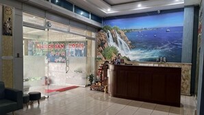 Uma área de recepção de hotel com um balcão, entrada por uma porta de vidro e um mural com paisagem costeira.