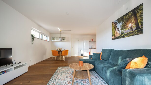 Un salon moderne avec un canapé, une table basse et un téléviseur. On y trouve également un coin repas avec des chaises et une table, ainsi qu’une cuisine à l’arrière-plan.