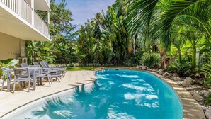 Una piscina con sillas de descanso, rodeada de plantas tropicales y una zona delimitada por una valla.