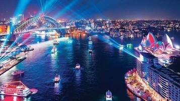Un paisaje urbano nocturno con un puente destacado, un teatro de ópera muy iluminado y un crucero amarrado en un puerto.