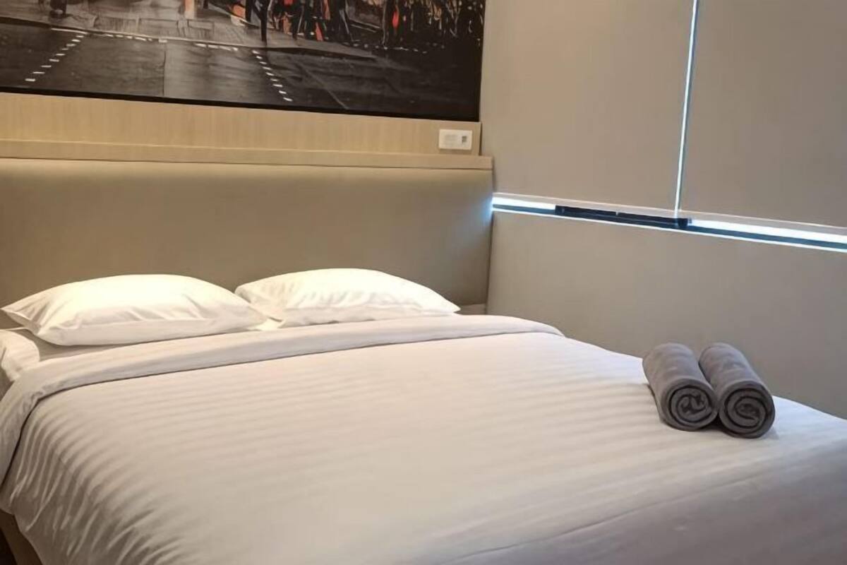 A neatly made bed with white linens and two rolled towels, a headboard, and a window with blinds.
