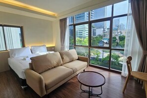Une chambre d’hôtel équipée d’un lit, d’un canapé, d’une table ronde et de grandes fenêtres donnant sur la ville.