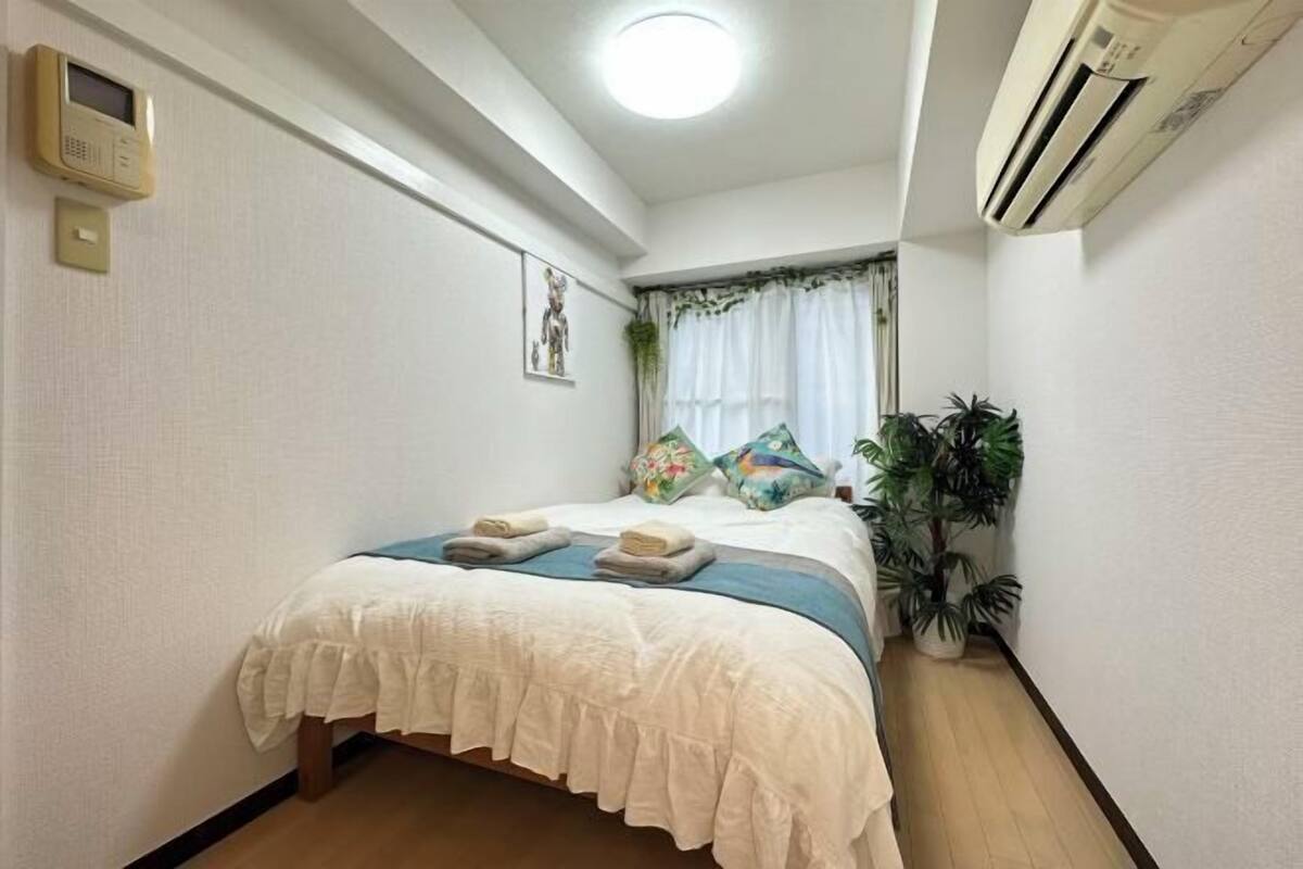 Una cama bien hecha con cubrecama blanco y detalles azules, una planta en la mesita de noche y un equipo de aire acondicionado fijado a la pared.