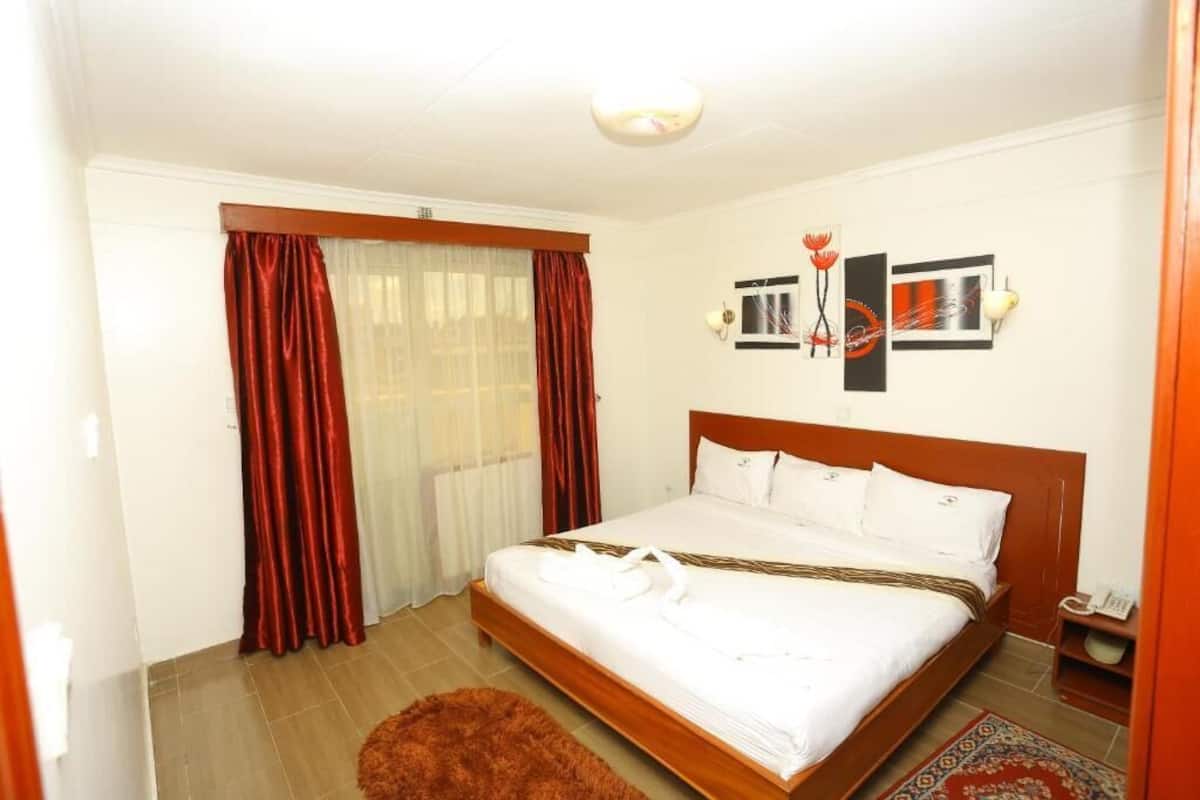 A hotel room with a wooden bed, white bedding, a red curtain, a white rug, and a small table with a lamp.
