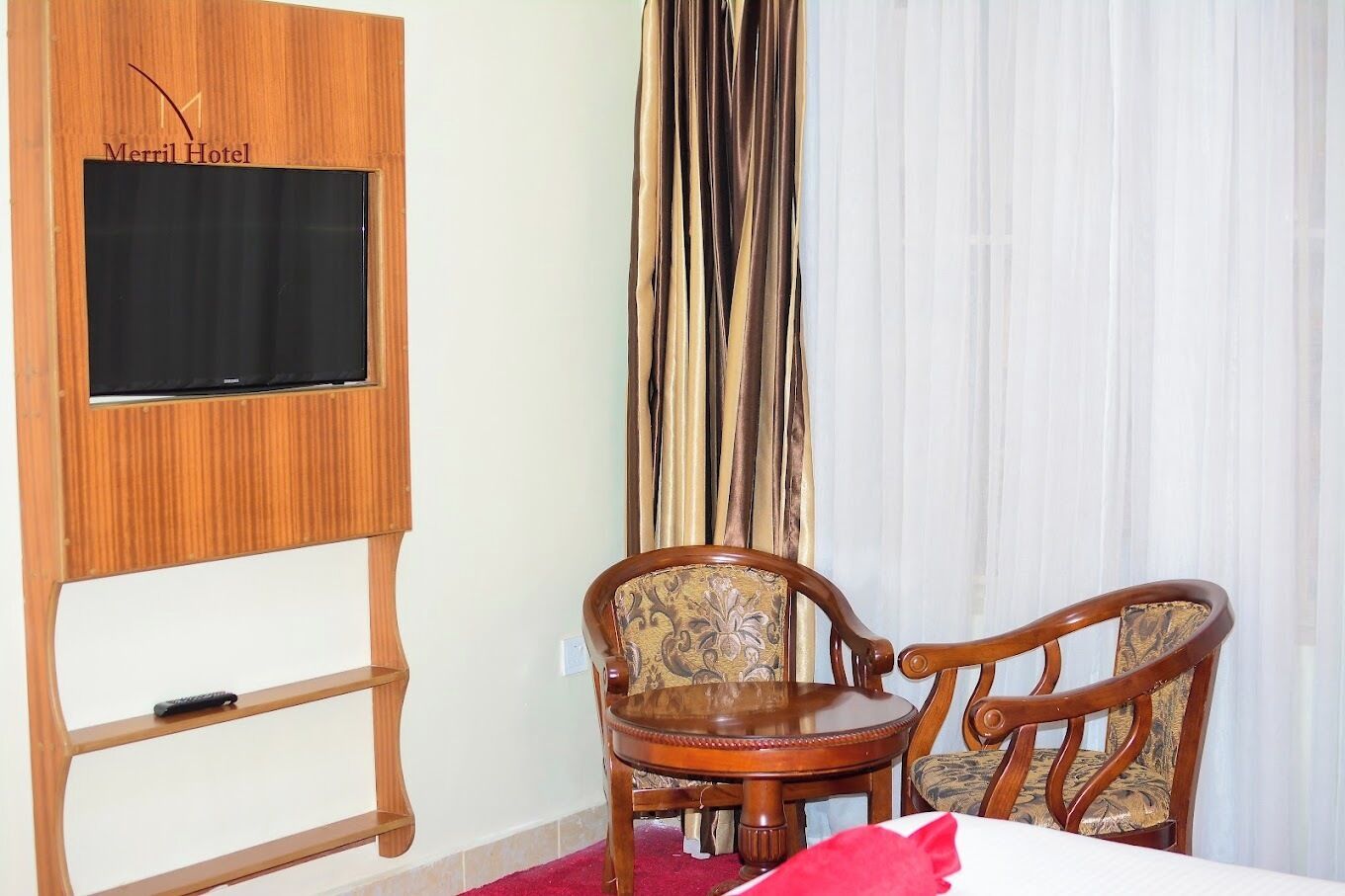 Ein Hotelzimmer mit einem HolztV-Schrank, einem Flachbildfernseher, zwei Holzstühlen, einem kleinen runden Tisch und einem roten Zierkissen.