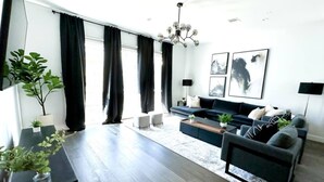 A modern living room with a dark sofa set, a coffee table, and a large window with black curtains. There are two potted plants on the windowsill and a chandelier hanging from the ceiling. - Luxe 3Bed Townhome in Heights with WiFi, Private Rooftop, Private Parking (Houston)