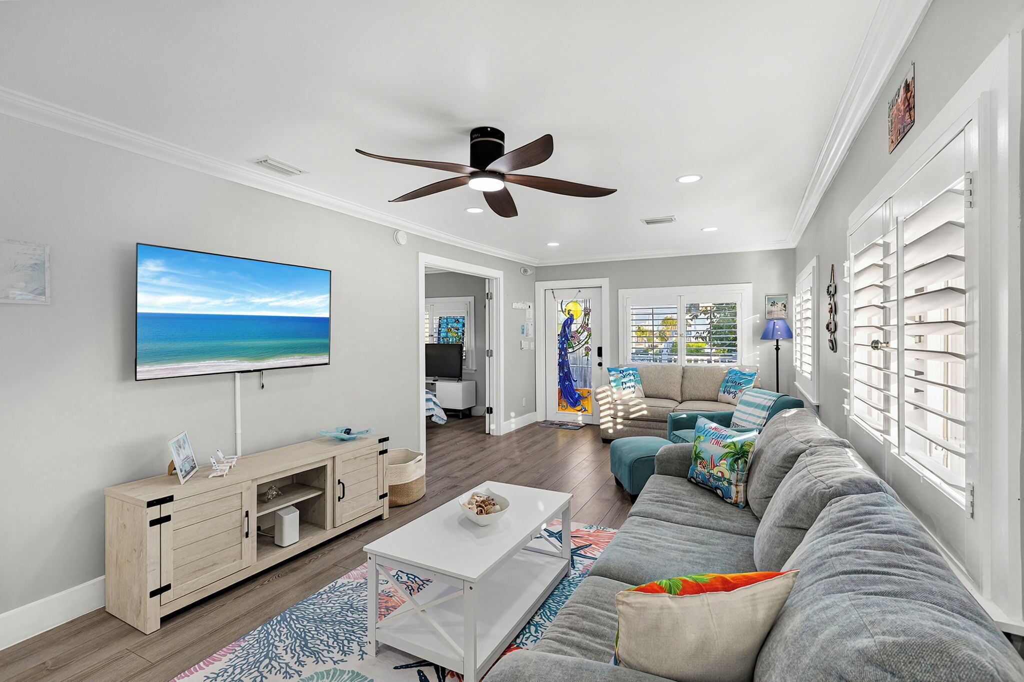 A modern living room with a flat-screen TV, a ceiling fan, and a large window with blinds.