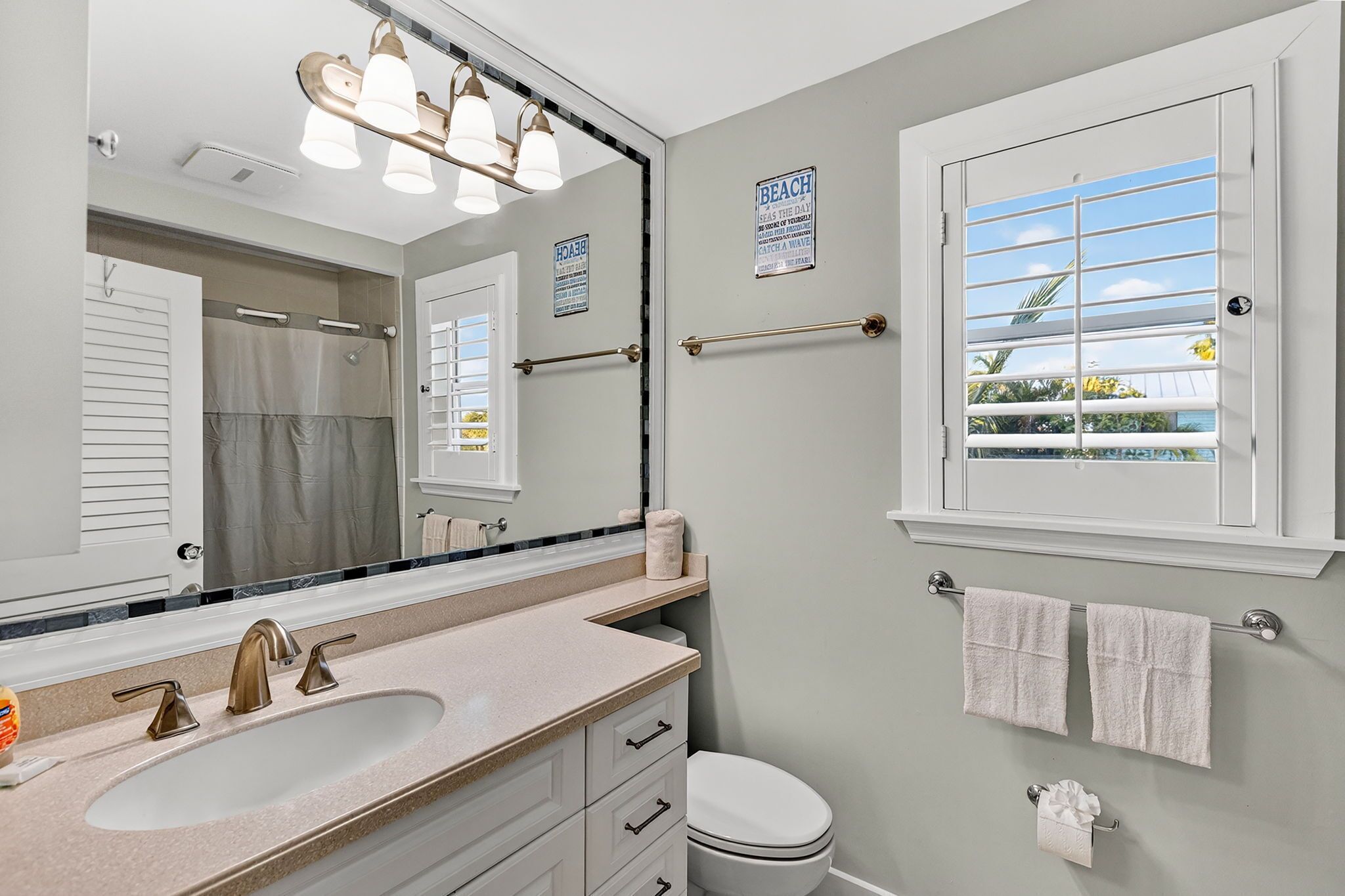 A bathroom with a large mirror, a sink with two faucets, a toilet, and a window with a view of the beach.