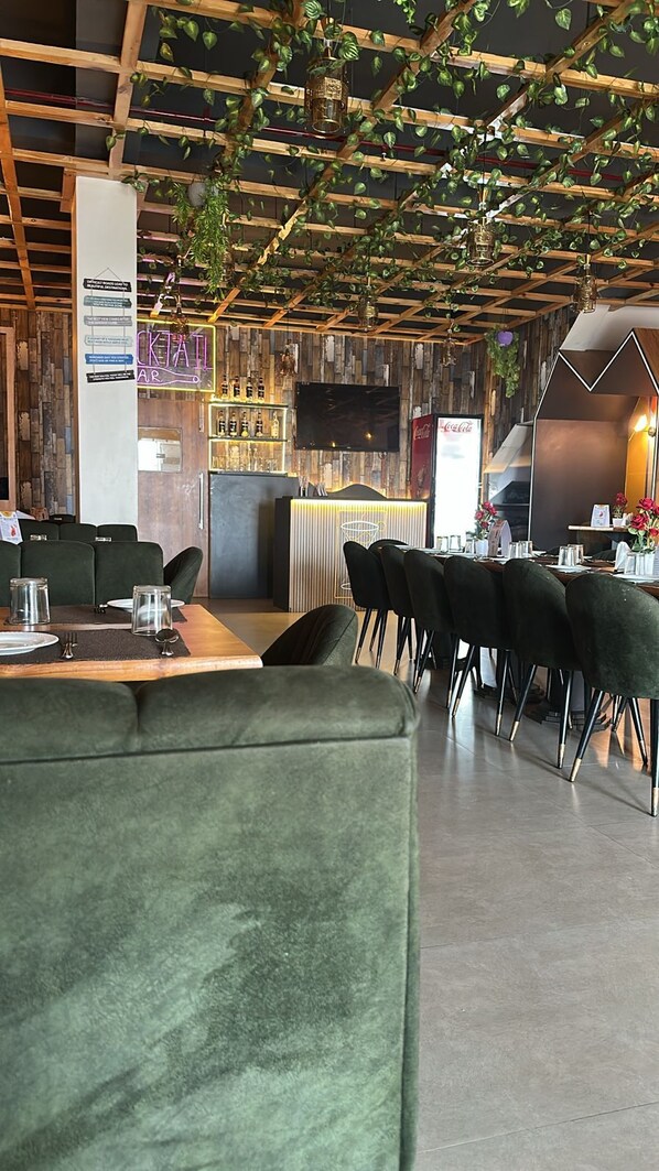 A modern restaurant with wooden ceiling beams, green upholstered seating, and a bar area with a neon sign. - The Divine Cloud Inn (Aligarh)