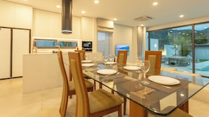A modern dining area with a glass table, wooden chairs, and a kitchen in the background.