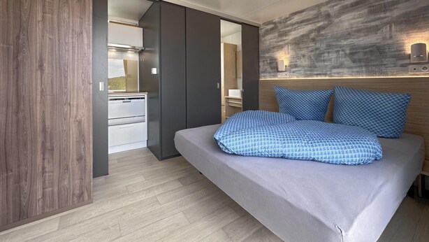 Une chambre moderne avec un lit, une armoire en bois et, au fond, un coin cuisine.