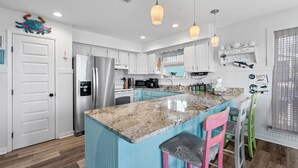 A modern kitchen with white cabinets, a large island with bar stools, stainless steel appliances, and pendant lighting. - Last Resort (Gulf Shores)
