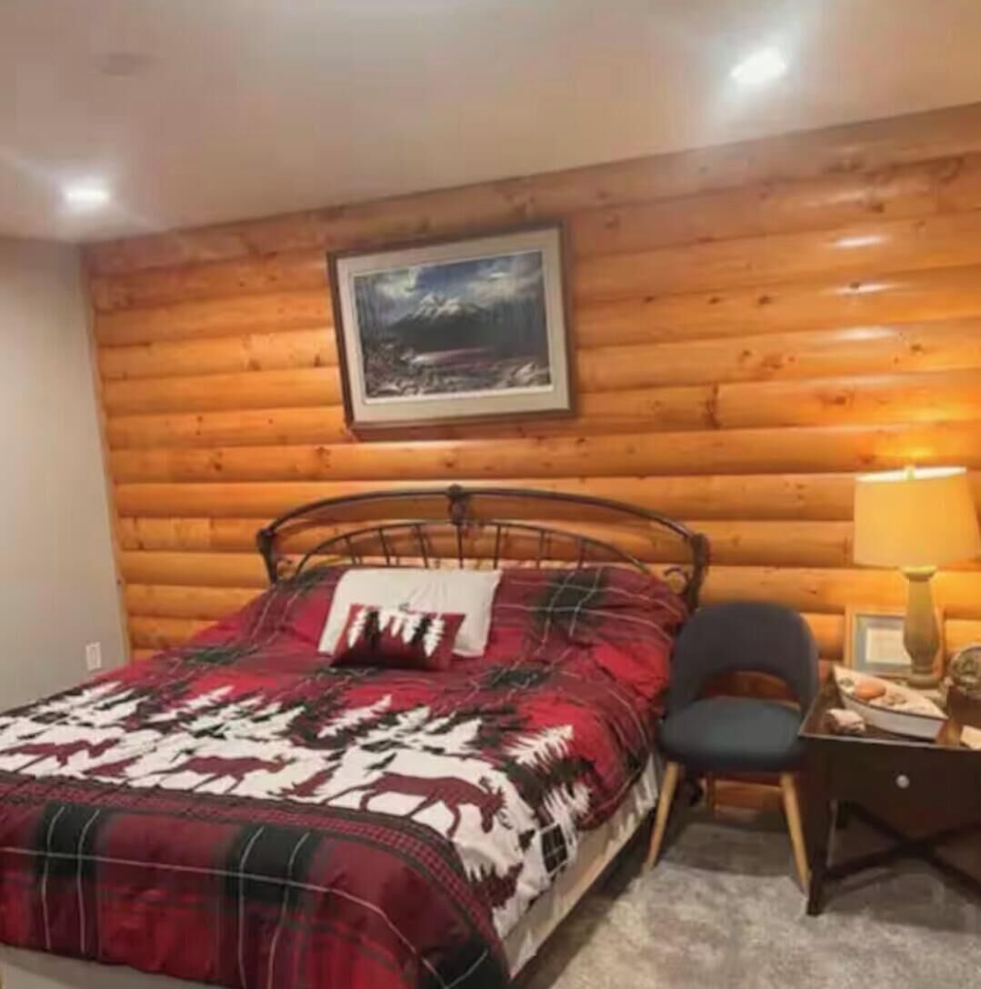 A bedroom with a wooden log wall, a bed with a red and white patterned blanket, a chair, a nightstand with a lamp, and a framed picture above the bed.