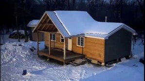 Een houten hut met een met sneeuw bedekte dak, een veranda en een klein bijgebouwt tuinhuis.