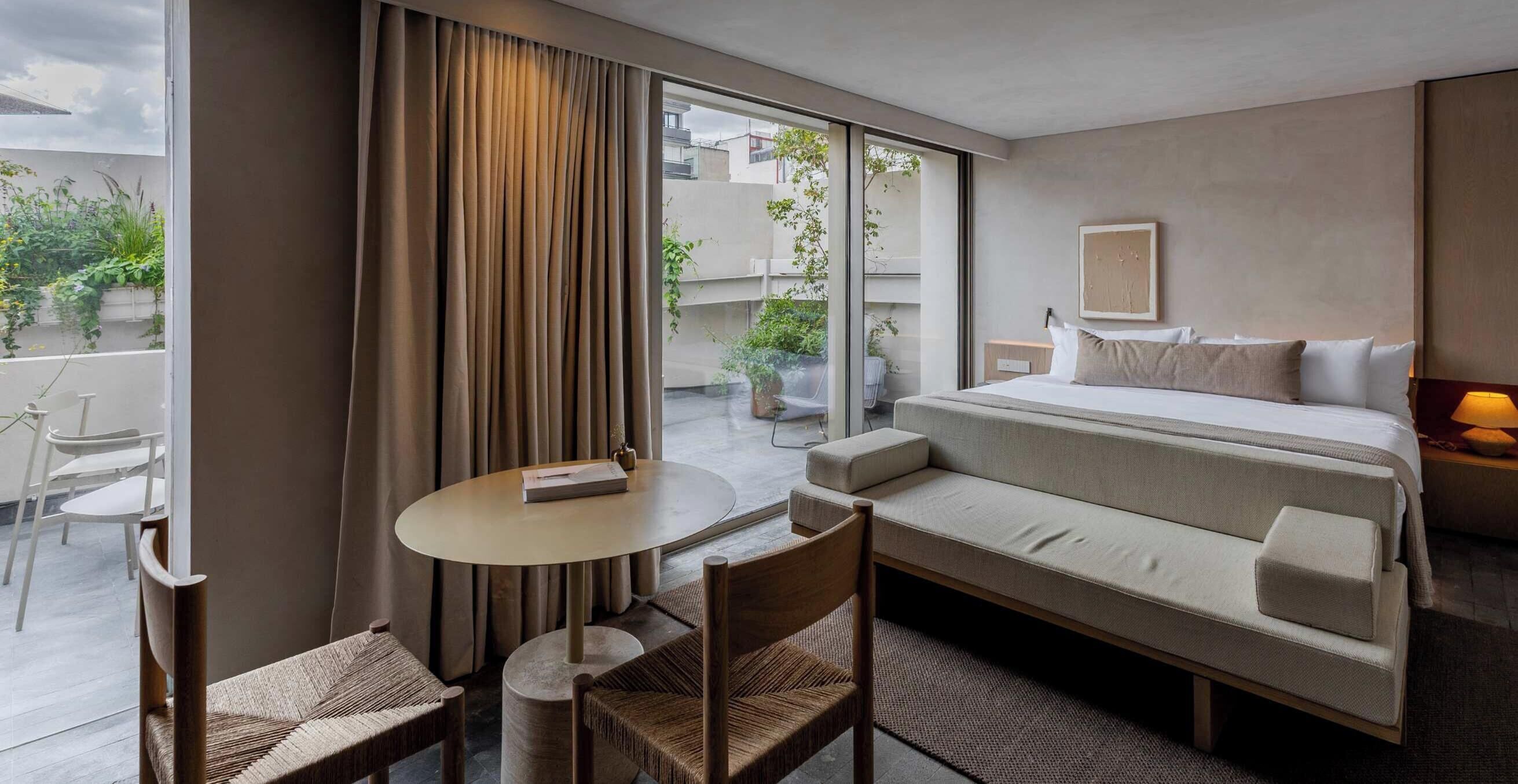 Ein Hotelzimmer mit einem Bett, einem runden Tisch, zwei Stühlen, einem Fenster mit Vorhängen und Blick ins Freie.