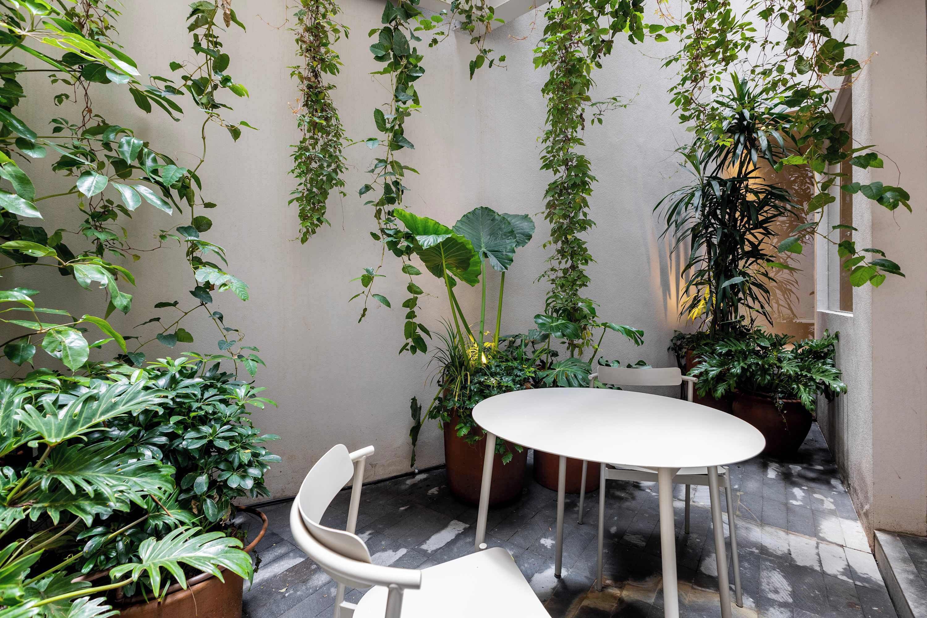 A small, enclosed patio area with a round table and a chair, surrounded by various potted plants and climbing vines.