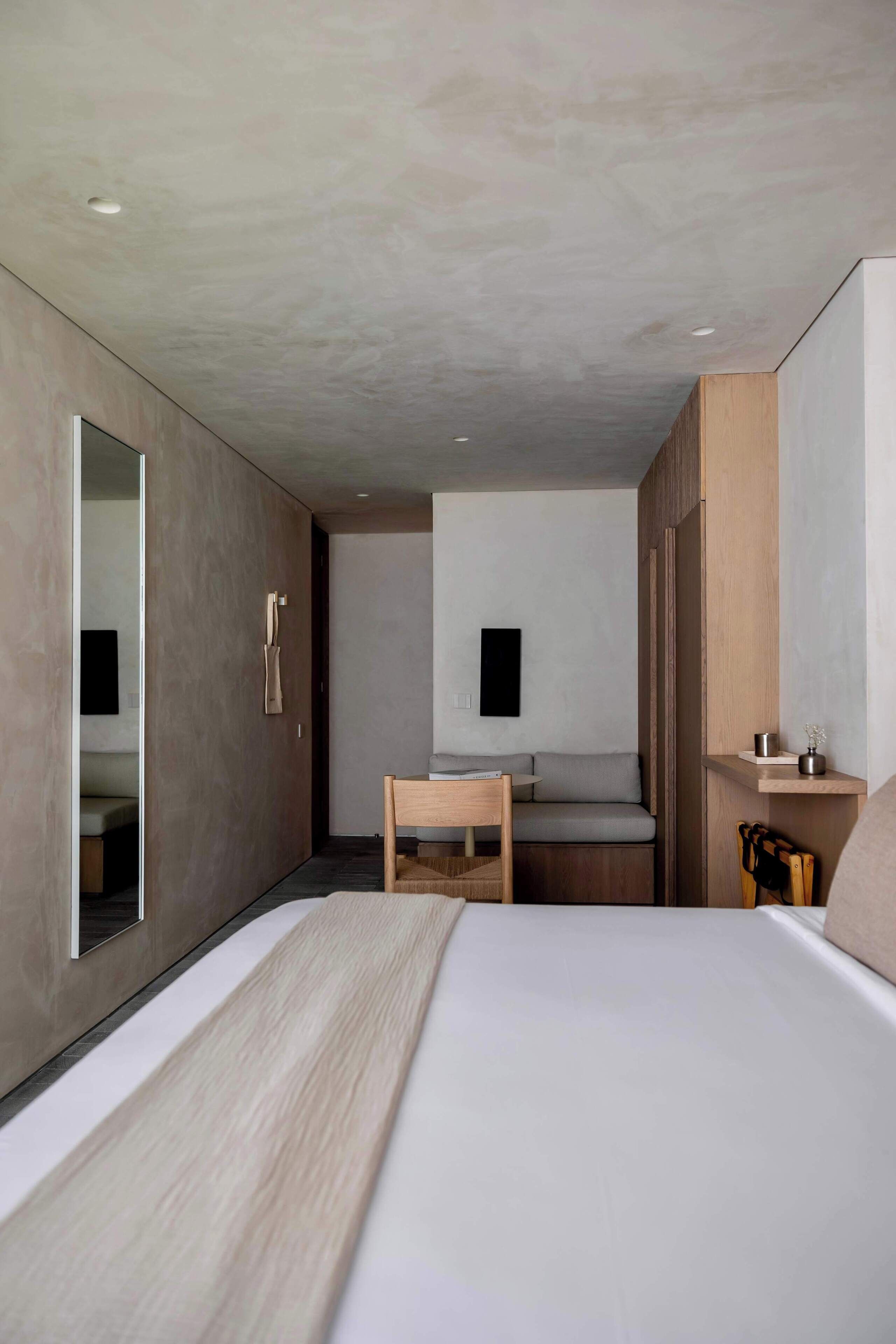A modern hotel room with a bed, a desk, a chair, a mirror, and a small shelf.