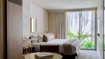 A modern hotel room with a large bed, a smaller bed, a dining table, and a view of greenery outside.